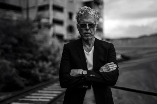 Black and white photo of a man standing on a boardwalk with sunglasses and arms crossed.