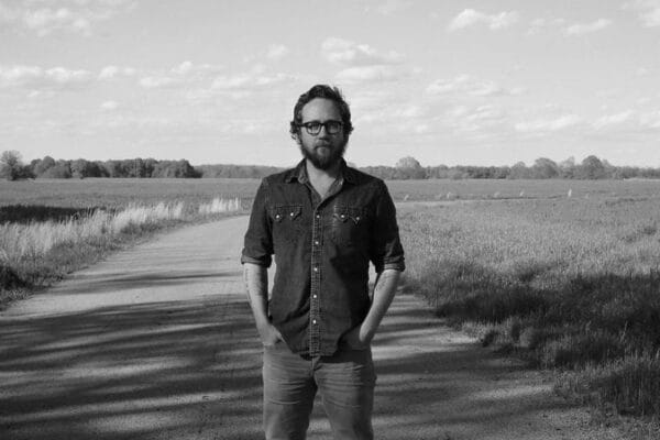 Over and over again with JPSK Justin Peter Kinkel-Schuster black and white photo man wearing glasses with hands in pockets standing on a dirt road between two fields