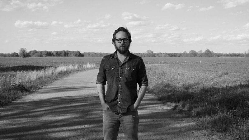 Justin Peter Kinkel-Schuster black and white photo man wearing glasses with hands in pockets standing on a dirt road between two fields