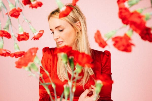 Emma Swift Rises Again: “The Resurrection Game” Emma Swift blonde woman wearing red looking sideways through red carnations