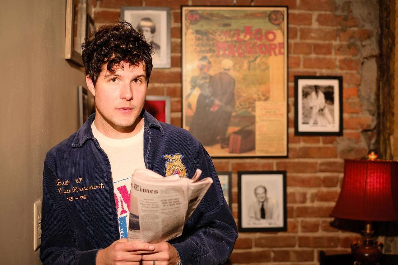 Brian Dunne standing in front of brick wall adorned with old pictures he's holding a newspaper