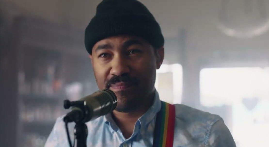 San Gabriel closeup wearing black beanie standing at microphone with visible rainbow stripe guitar strap