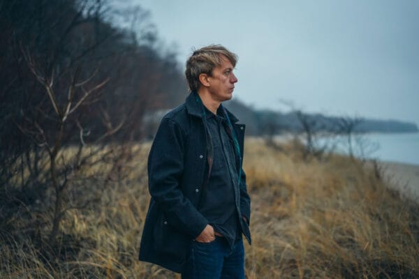 Steve Gunn’s Music for Writers Offers a Quiet Revolution in Sound Man with sandy blonde hair wearing a dark blue coat with hands in pocket standing brush looking out over water