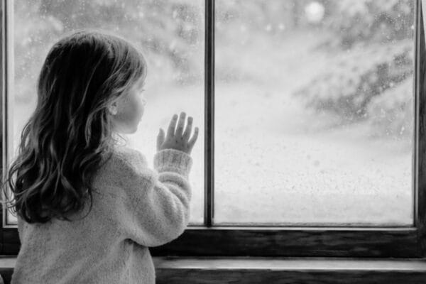 A small child stands at a window, seen from behind, with one hand resting on the glass as snow falls outside.