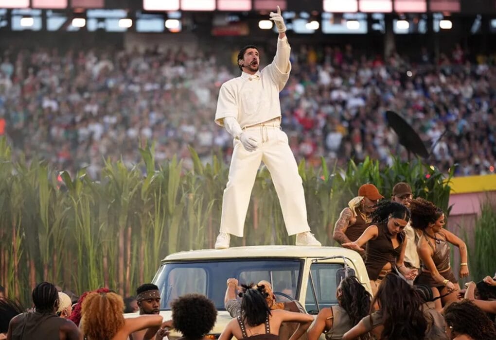 Bad Bunny stands on the roof of a white SUV in a sugarcane field set, wearing an all-white outfit and pointing upward as dancers perform around him in a packed stadium.