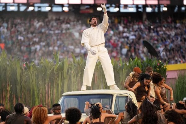 Bad Bunny stands on the roof of a white SUV in a sugarcane field set, wearing an all-white outfit and pointing upward as dancers perform around him in a packed stadium.