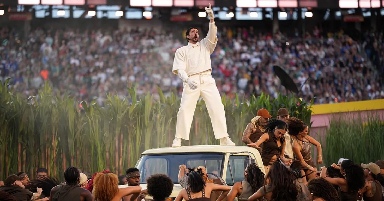 Bad Bunny stands on the roof of a white SUV in a sugarcane field set, wearing an all-white outfit and pointing upward as dancers perform around him in a packed stadium.