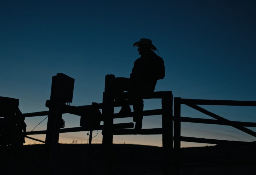 Caleb Hearn wearing a cowboy hat and sitting on a fence against a dark sky sunset background