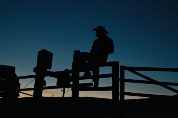 Caleb Hearn wearing a cowboy hat and sitting on a fence against a dark sky sunset background