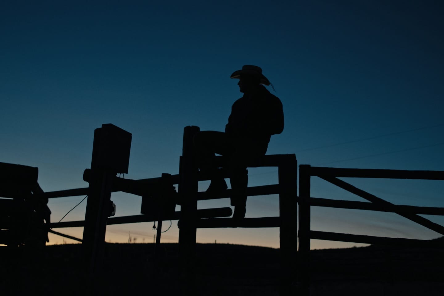 Caleb Hearn wearing a cowboy hat and sitting on a fence against a dark sky sunset background